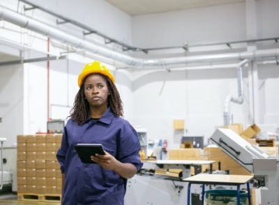 Serious African American female worker in protective uniform walking to workplace on plant floor, holding tablet and case with tools. Vertical shot. Women in industry concept