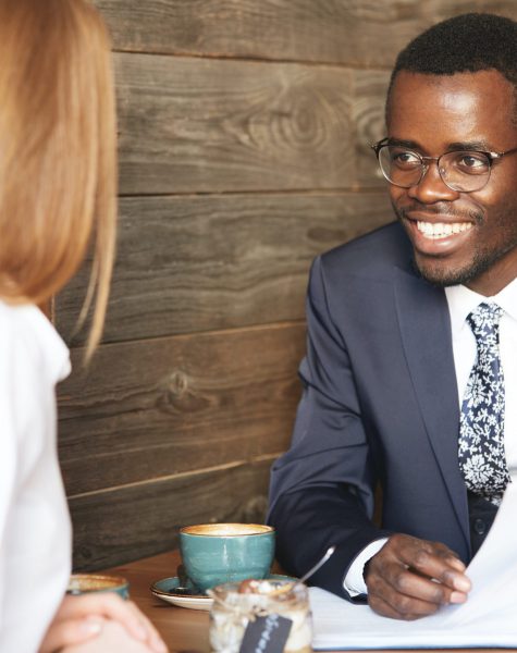 Successful negotiations. Smiling cheerful African American entrepreneur wearing glasses and formal suit, having a meeting with his redhead Caucasian female partner in white shirt during lunch break